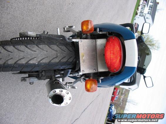 Rear of a blue and white motorcycle showing the tail light, turn signals, exhaust, and tire. Supermotors.net logo visible