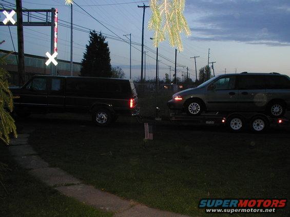 Black SUV towing a gray minivan on a dual-axle trailer at dusk