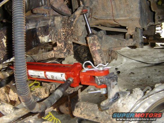 Hydraulic jack attached to vehicle suspension with cotter pins. Supermotors.net logo visible