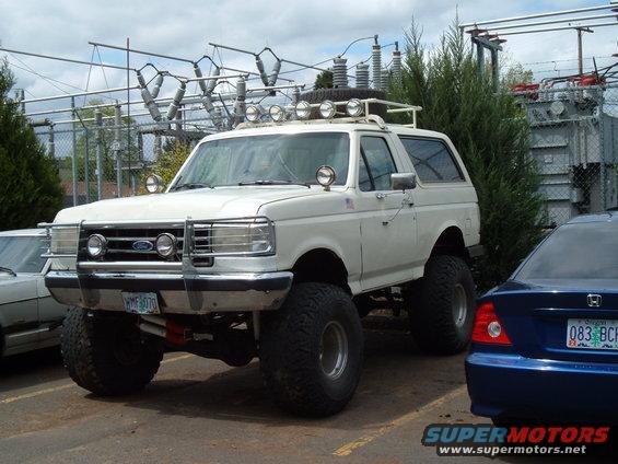 White 1989 Ford Bronco lifted with off-road lights on roof rack and bull bar. Blue sedan parked to the right