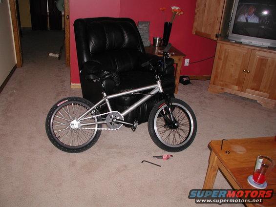 Silver BMX bike parked indoors next to a black leather chair. Tools and a drink are on the floor nearby