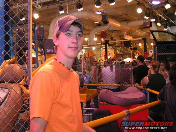 Young man in orange t-shirt and cap, with a blurred background of people and what appears to be a car show