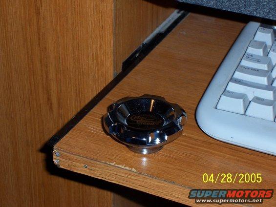 Chrome gas cap with "Max Speed" text on a wooden surface next to a keyboard