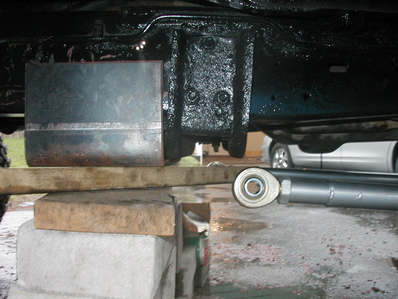 close-up of metal plate welded to vehicle frame, supported by blocks and wood, with a suspension component visible