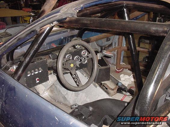 Interior of a modified car with a roll cage, racing steering wheel, and custom dashboard. The car appears to be stripped down for racing