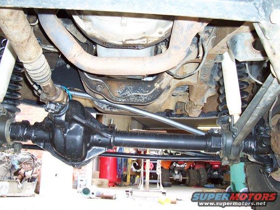 Underside view of a truck's rear axle, driveshaft, and suspension components. Other vehicles are visible in the background