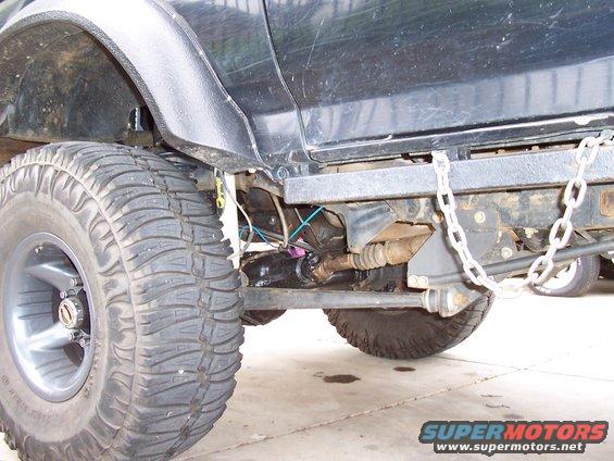 Black truck's front axle and suspension with large off-road tire and silver rim. Chain attached to frame