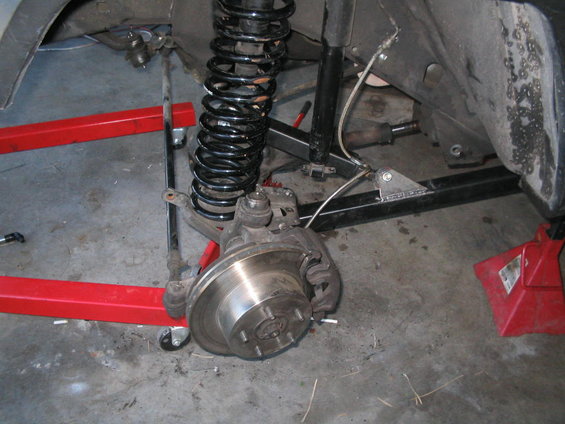 Front suspension assembly with coil spring, shock absorber, brake rotor, and caliper. Red jack stands and a red creeper are visible