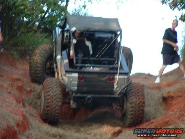 Off-road vehicle with large tires climbing a dirt hill. Roll cage visible. Person in background