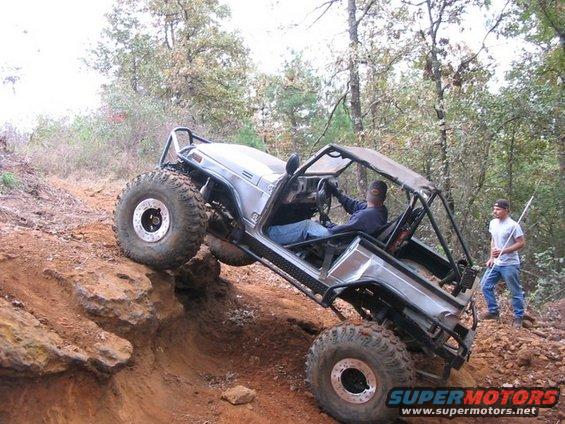 Modified off-road vehicle with large tires climbing a steep dirt incline, two people visible