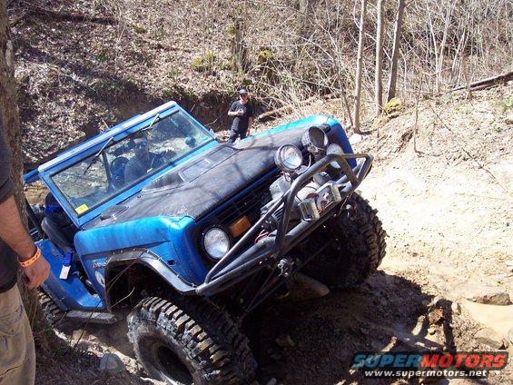 Blue first-generation Ford Bronco with a roll cage, off-roading on a muddy trail