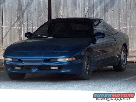 Dark blue coupe parked in front of a wooden fence. The car has a sporty design with a low profile and tinted windows