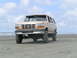 1988 Ford C-250 (Centurion Conversions) - Tan Ford Bronco with aftermarket wheels and tires parked on a sandy beach with the ocean in the background.