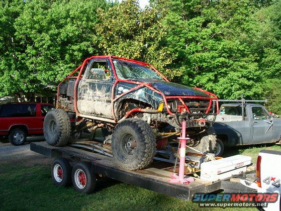 Muddy truck with red roll cage on a trailer, parked next to a red SUV and a gray pickup truck