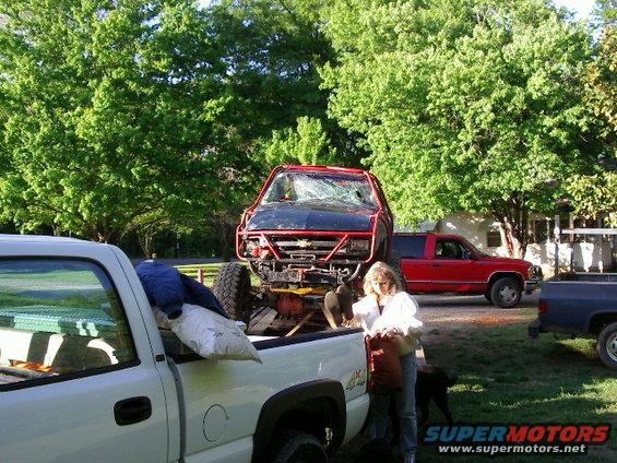 Three trucks outdoors, one black truck with red roll cage loaded onto a white pickup bed, a red SUV parked behind, and a dark blue truck to the right