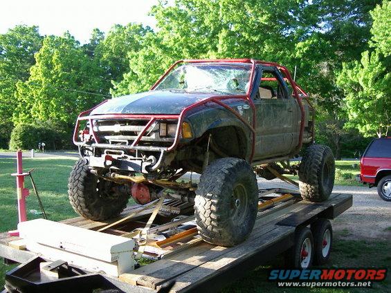 Off-road truck with roll cage and large tires on a trailer, red SUV in background