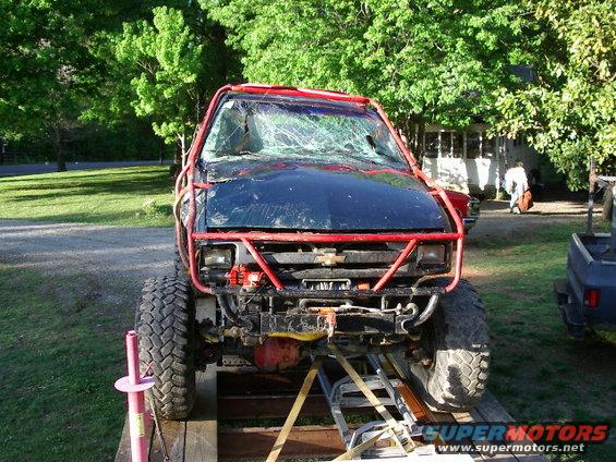 Black off-road truck with red roll cage and cracked windshield, front view, on a trailer