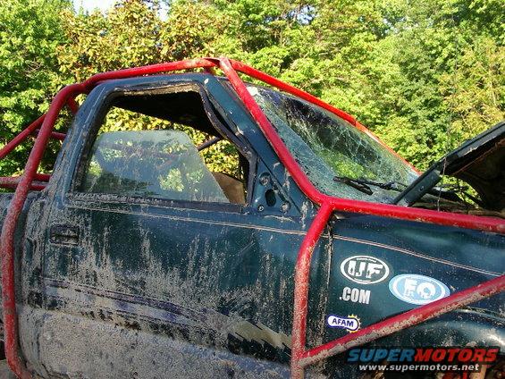 Mud-splattered green truck with red roll cage, broken windshield, and stickers on the door