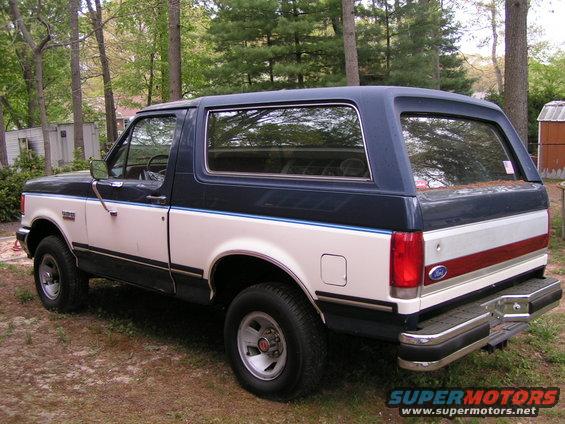 Two-tone white and blue 1987 Ford Bronco parked on dirt with trees in background