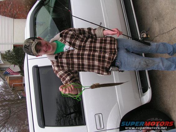 Young person in plaid shirt and jeans holding fishing rod and knife, standing next to white SUV