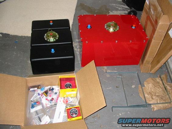 Three black fuel tanks stacked, one red fuel tank, and a box of gauges and parts on a concrete floor