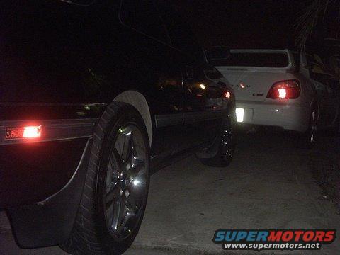 Rear view of a white Subaru WRX and a dark car parked at night. "SUPERMOTORS" logo visible