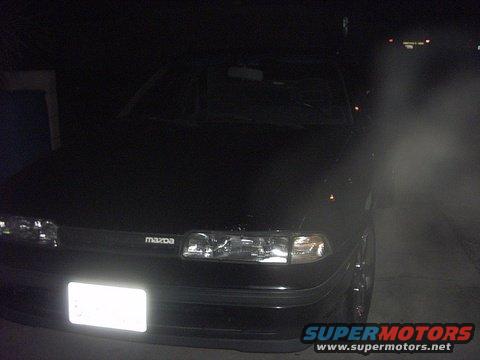 Front view of a black 1989 Mazda MX-6 with headlights on