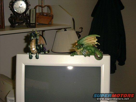 Two green dragon figurines perched on the edge of a white computer monitor, with a clock and basket on a shelf above