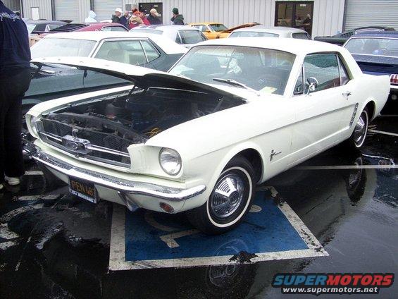 White 1965 Ford Mustang coupe with hood open, parked on a blue handicapped spot. Other cars visible in background