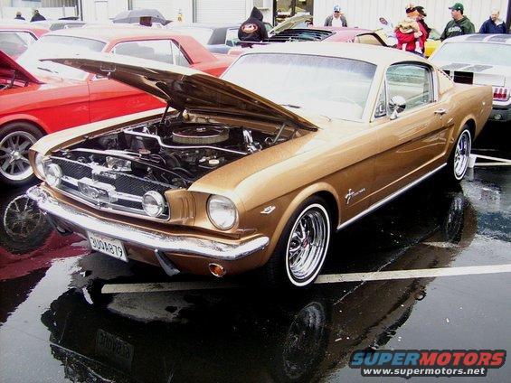 Gold 1965 Ford Mustang coupe with hood open, chrome wheels, and whitewall tires parked on wet asphalt