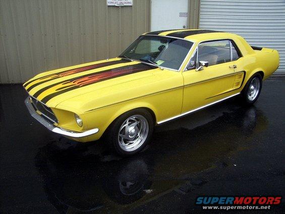 Yellow 1967 Ford Mustang coupe with black and orange racing stripes on the hood and roof, parked on a wet surface
