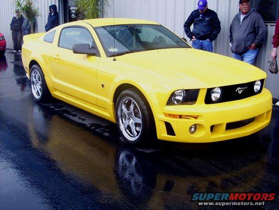 Yellow Ford Mustang parked on wet asphalt. People stand in the background