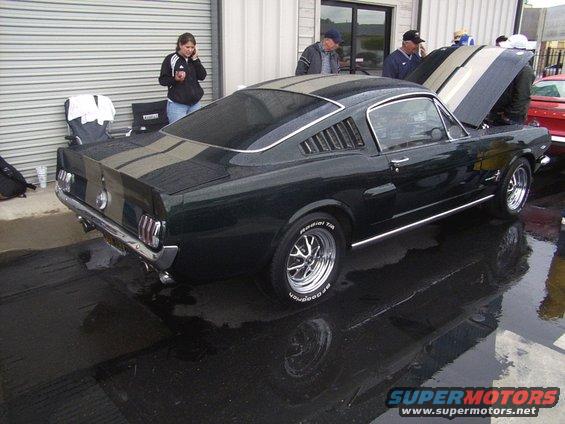 Dark green 1965 Ford Mustang with silver racing stripes, chrome wheels, and louvers on the rear quarter windows
