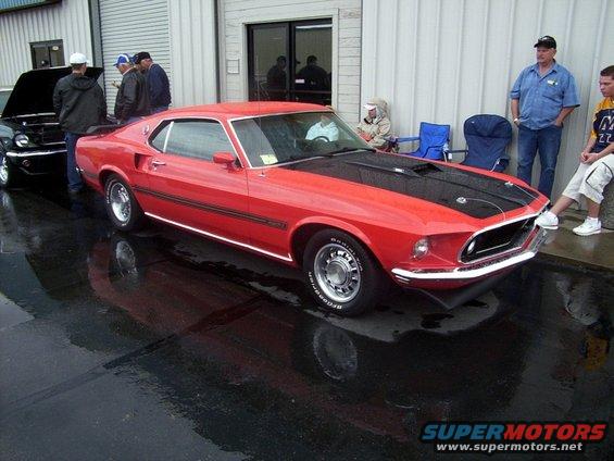Red 1969 Ford Mustang fastback with black hood stripes parked on wet asphalt