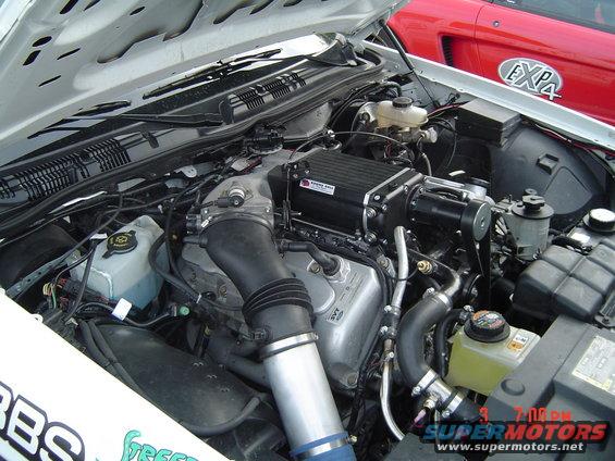 Close-up of a supercharged V8 engine bay, with a red car visible in the background