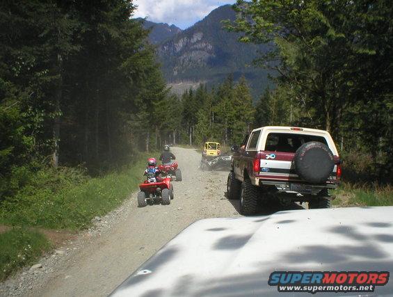 Maroon and white 1978 Ford Bronco with spare tire on rear, driving on a dirt road
