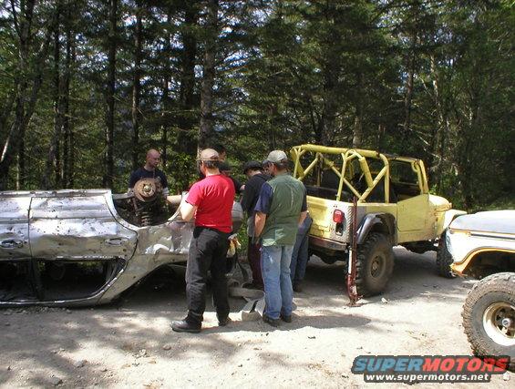 People gathered around a severely damaged car body and a yellow Jeep with a roll cage. Another vehicle is partially visible