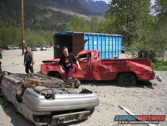Guy in a t-shirt sits on a wrecked red pickup next to an overturned car. Blue dumpster in background