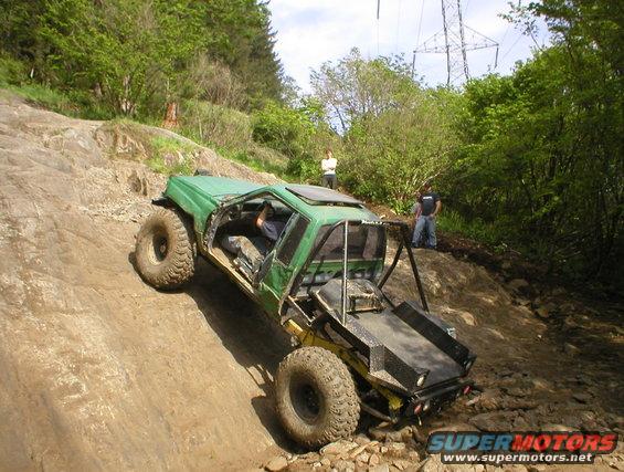 Green off-road vehicle with large tires climbing a rocky incline, roll cage visible