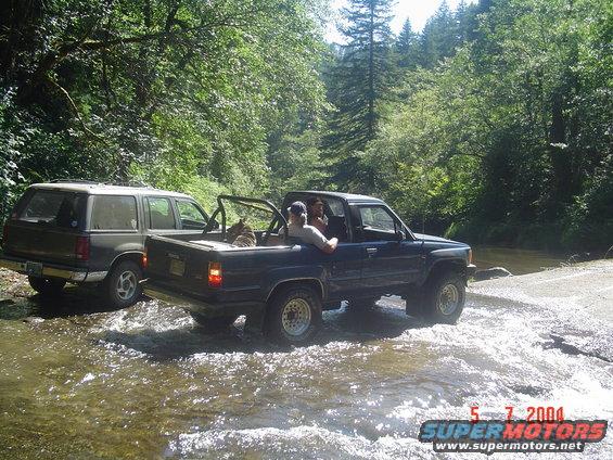 Dark blue pickup truck and a dark SUV driving through a stream in a wooded area