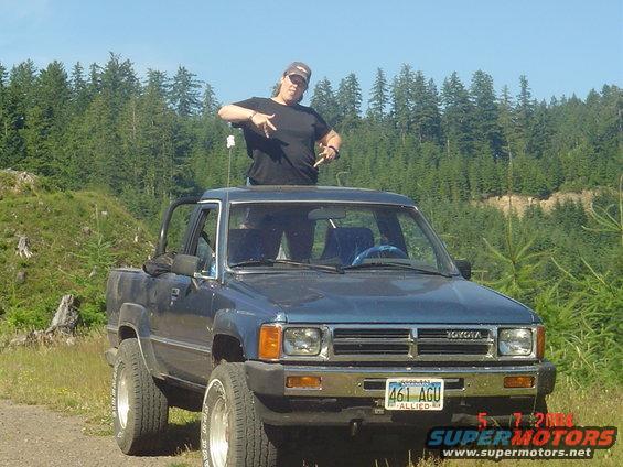 Blue 1984 Toyota pickup truck, front view, with person standing through sunroof