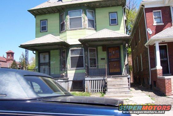 Dark blue car hood and roofline in foreground, with a green house and a brick house in the background