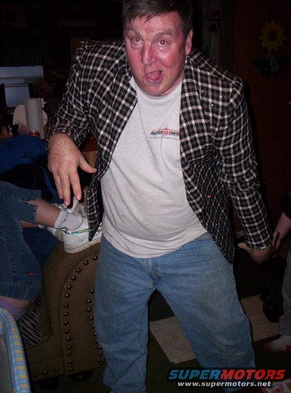 Man in a plaid jacket and jeans makes a funny face, with other people visible in the background