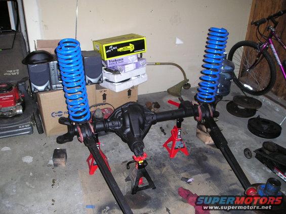 Vehicle axle assembly with blue springs on jack stands, Yukon box and lawnmower in background