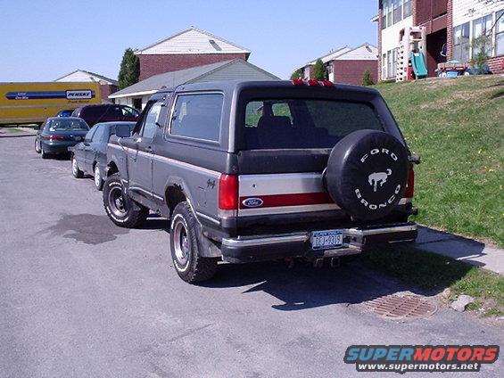Rear view of a 1987 Ford Bronco parked on a street, with a spare tire on the back. Other cars and a Pensky truck are visible