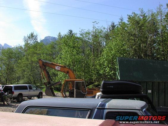 Excavator parked next to a silver SUV and other vehicles, with trees and mountains in the background