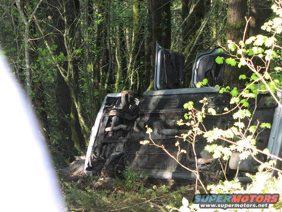 Overturned vehicle, possibly a Ford Bronco, lying on its side in a wooded area. Doors are open