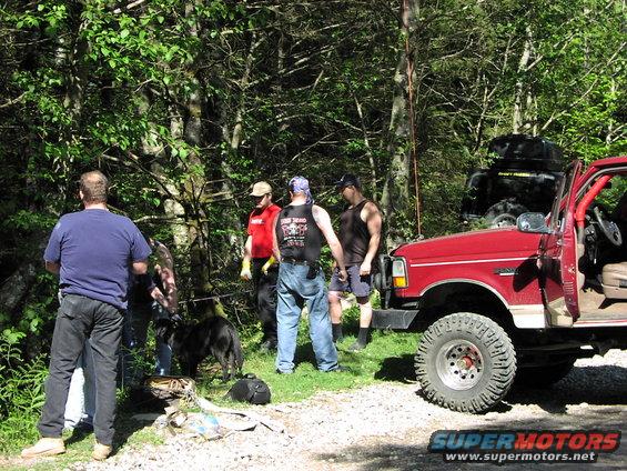 red off-road truck with people standing around it in a wooded area, black SUV in background