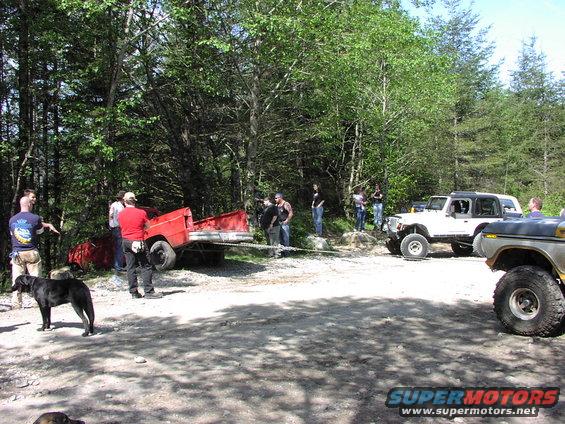 People gathered around a red trailer and several off-road vehicles in a wooded area