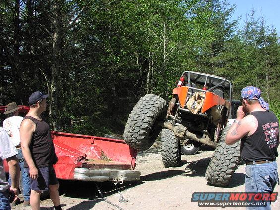 Orange off-road buggy with large tires is stuck, being pulled by a red pickup truck. People watch